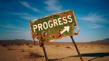 A heavily rusted and damaged sign reading "PROGRESS" with an arrow stands tilted in a barren desert landscape under blue sky.