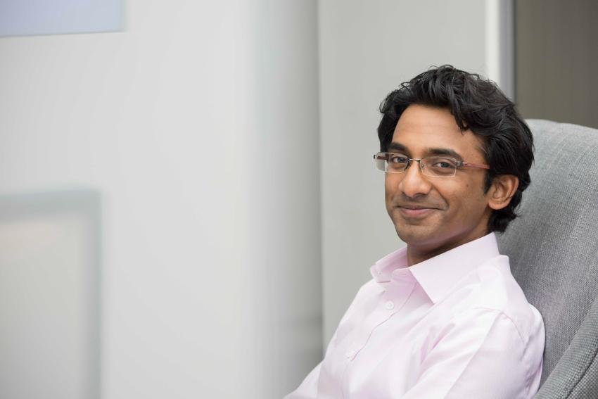 A person with glasses sits in a chair, wearing a light pink shirt, and looks towards the camera with a slight smile.