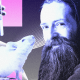 A man with a long beard sits next to a white lab mouse while a gloved hand holds a test tube in the background with abstract purple and pink shapes.