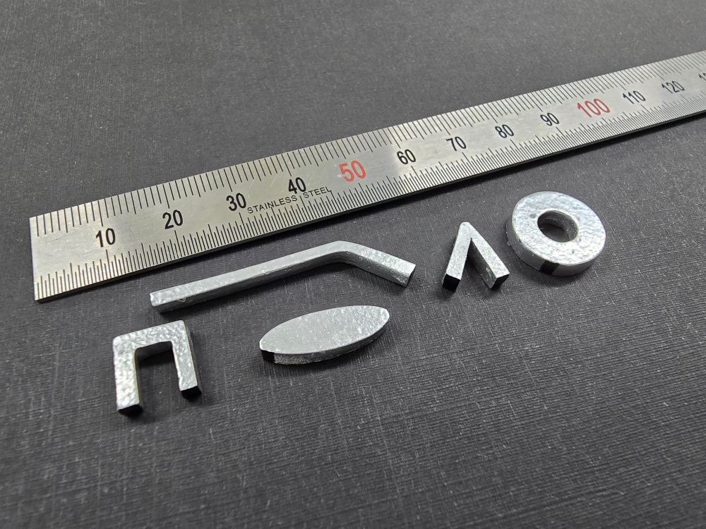 A sleek stainless steel ruler lies alongside five metallic pieces, meticulously arranged in intriguing shapes on a dark surface, reminiscent of avant-garde wearables.